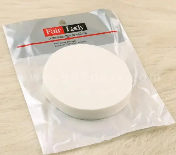 Éponge de Maquillage Blanche Fair Lady (1 Outil pour un Teint Parfait) Éponge de maquillage blanche ronde Fair Lady en latex NBR, dans son emballage plastique transparent, posée sur une fausse fourrure beige.