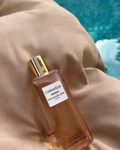 Flacon Caramella Emmanuelle Jane 265 ml doré, posé sur tissu beige près d'une eau bleutée, éclat chic et douceur gourmande.