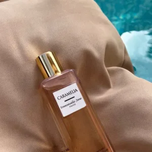 Flacon Caramella Emmanuelle Jane 265 ml doré, posé sur tissu beige près d'une eau bleutée, éclat chic et douceur gourmande.