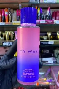 Main gantée présentant le flacon dégradé rose et bleu de la brume My Way pour corps et cheveux, sur une étagère de produits.