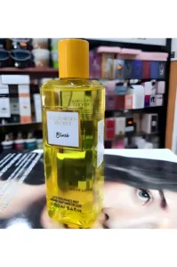 Flacon jaune transparent brume Blush Victoria's Secret, 250ml, posé sur un magazine devant un étalage de parfums colorés.