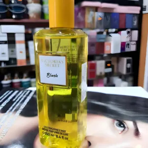 Flacon jaune transparent brume Blush Victoria's Secret, 250ml, posé sur un magazine devant un étalage de parfums colorés.