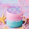 Pot de gommage corporel Maribelle Sugar Scrub Mermaid Pastel, bicolore rose et bleu, posé sur une surface de sable et de perles.