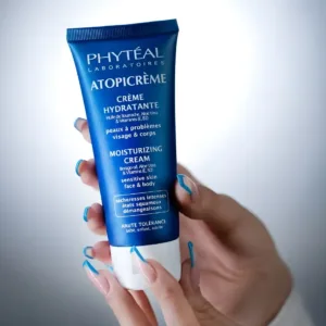 Tube bleu de crème hydratante Phyteal Atopicrème pour peaux sensibles, tenu par des mains aux ongles soignés sur fond clair.