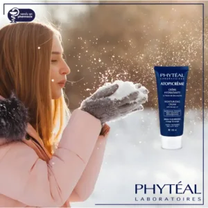 Jeune femme en extérieur hivernal soufflant de la neige, à côté d'un tube de crème Phyteal Atopicrème sur fond de paysage.