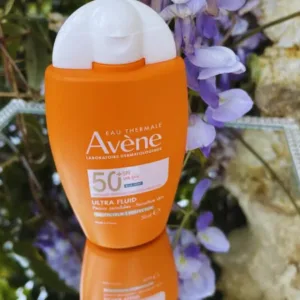Flacon orange Avène Solaire Ultra Fluid SPF 50+ posé sur un miroir, avec un arrière-plan flou de glycine mauve en fleurs.