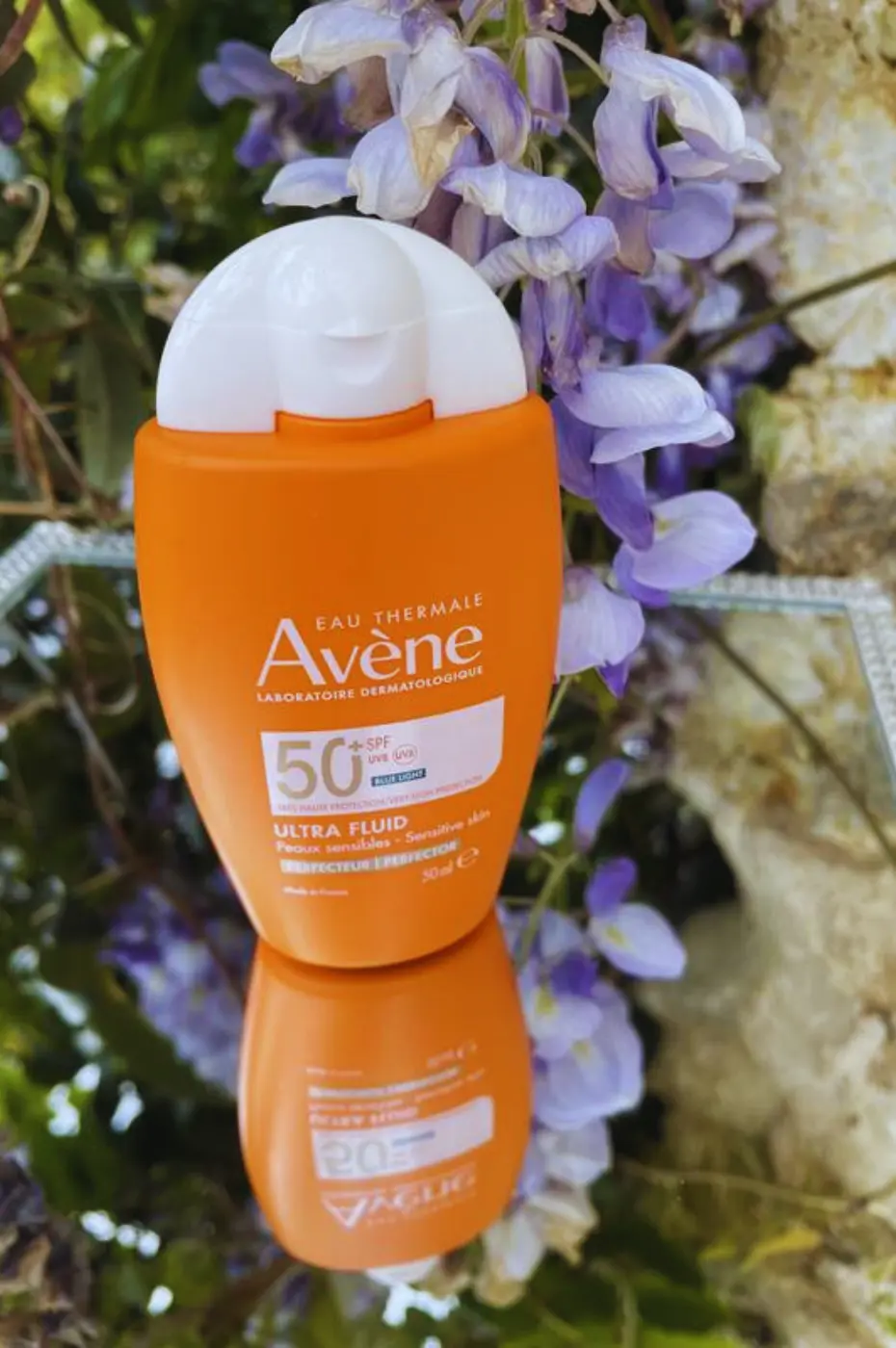 Avène Solaire Ultra Fluid SPF 50+ : Protection Visage Naturelle Flacon orange Avène Solaire Ultra Fluid SPF 50+ posé sur un miroir, avec un arrière-plan flou de glycine mauve en fleurs.