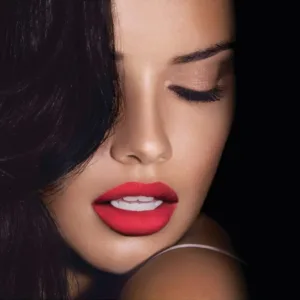 Gros plan sur un mannequin aux cheveux noirs, avec un rouge à lèvres Maybelline Mat 965 éclatant et des paupières fermées nude.
