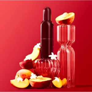 Flacon de parfum Dark Red entouré de tranches de pêches juteuses et de fleurs blanches. Mise en scène fruitée sur fond rouge.