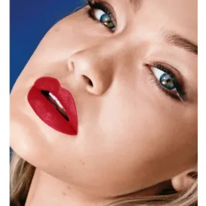 Mannequin blond portant le rouge à lèvres Maybelline 965. Rouge mat vif sur les lèvres, yeux charbonneux, fond bleu.