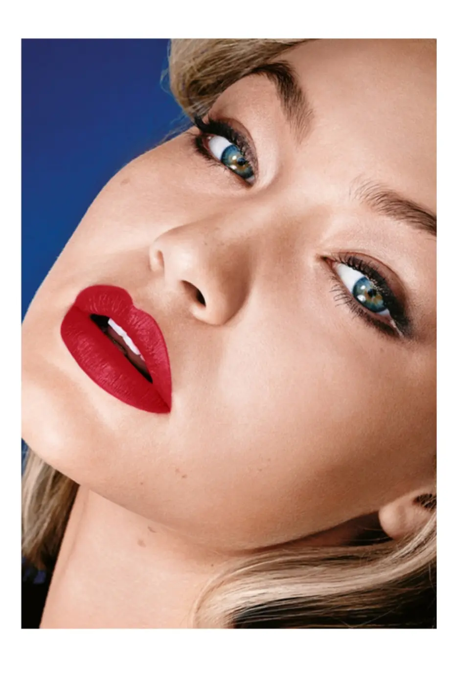 Glamour Intense Mannequin blond portant le rouge à lèvres Maybelline 965. Rouge mat vif sur les lèvres, yeux charbonneux, fond bleu.