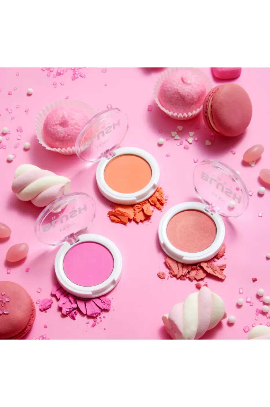 HEAN BLUSH BABY – Trio Gourmand de Couleurs Poudrées Trois boîtiers de blush HEAN BLUSH BABY (rose vif, corail, rose pêche) entourés de macarons, guimauves et bonbons sur un fond rose.