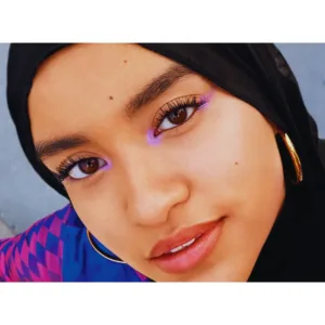 Portrait d'une jeune femme portant un hijab noir et un eye-liner flottant lilas vif sur les yeux. Grosses boucles d'oreilles dorées.