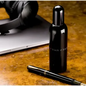 Bouteille noire du parfum Colour Me Black à côté d'un stylo-plume noir, d'un ordinateur portable argenté et d'un casque audio.