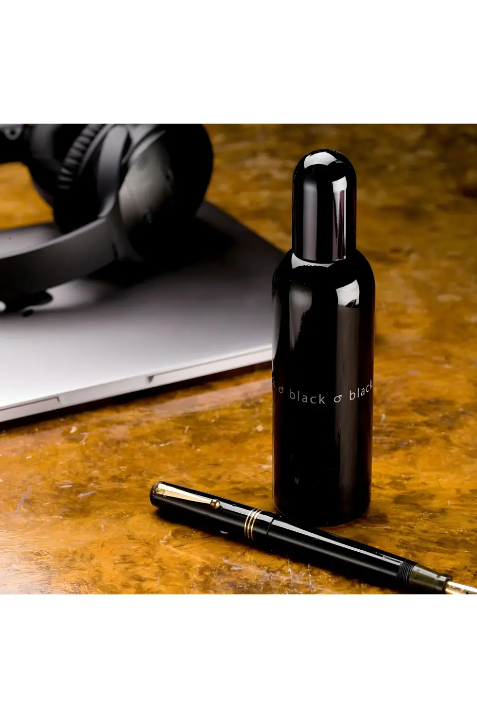 L’Essence de l’Homme Moderne Bouteille noire du parfum Colour Me Black à côté d'un stylo-plume noir, d'un ordinateur portable argenté et d'un casque audio.