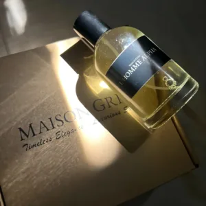 Flacon de parfum L'Homme Alpha Maison Griri sur boîte dorée, lumière du soleil. Parfum masculin élégant.
