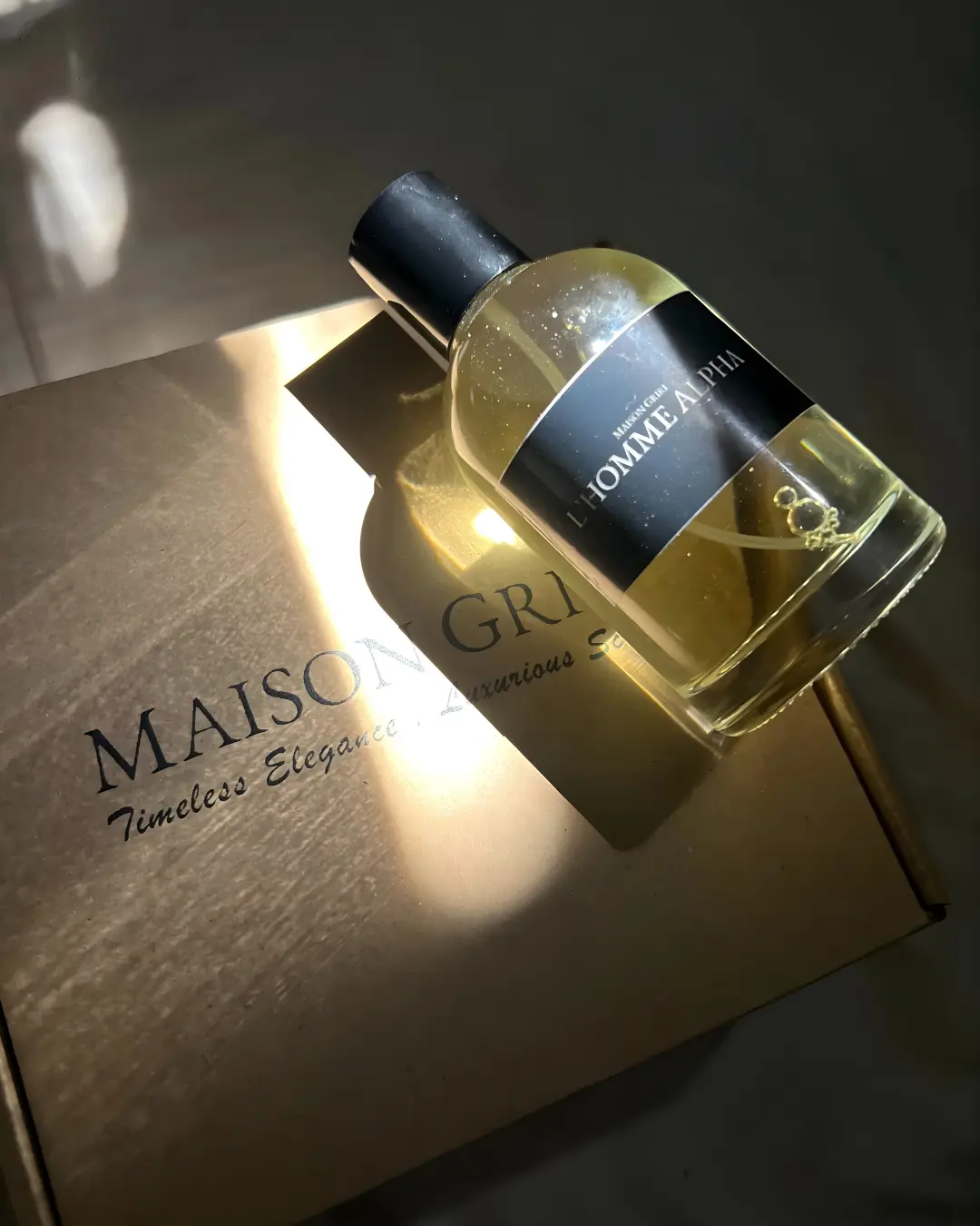 L'Homme Alpha Flacon de parfum L'Homme Alpha Maison Griri sur boîte dorée, lumière du soleil. Parfum masculin élégant.