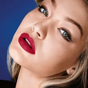 Portrait en gros plan d'une femme blonde aux yeux bleus intenses, inclinant la tête, portant le rouge à lèvres Maybelline Matte N° 960 Red Sunset sur un fond bleu nuit.