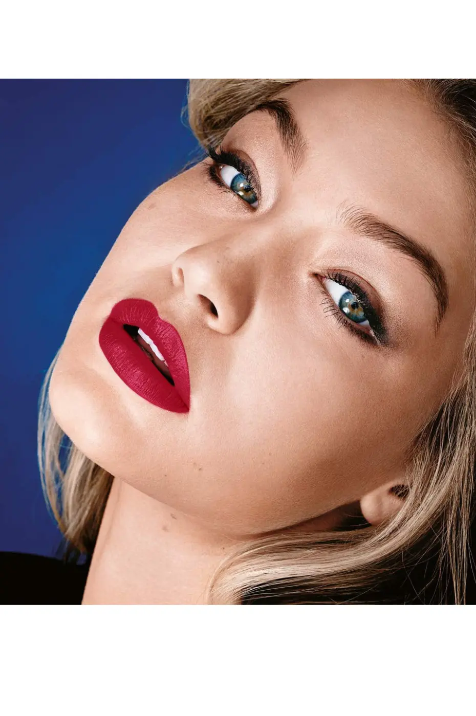 Regard Magnétique : Red Sunset et yeux bleus intenses Portrait en gros plan d'une femme blonde aux yeux bleus intenses, inclinant la tête, portant le rouge à lèvres Maybelline Matte N° 960 Red Sunset sur un fond bleu nuit.
