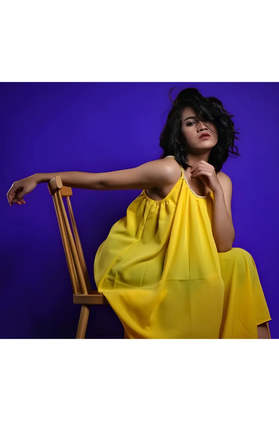 Révélez votre style Portrait d'une jeune femme portant une robe jaune sur fond indigo, assise sur une chaise en bois, exprimant sa confiance.