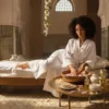 Une femme aux cheveux bouclés en peignoir blanc, assise dans un spa d'inspiration tunisienne, trempe délicatement ses doigts dans un bol en bois rempli d'eau et de pétales de rose. Un plateau doré avec des huiles et des crèmes repose à côté d'elle, sous une douce lumière matinale.