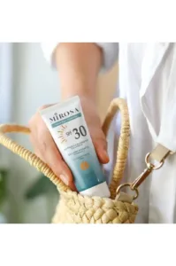 Main tenant un tube de crème solaire MIROSA SPF 30 sortant d'un sac en osier, sur fond de chemise blanche lumineuse.