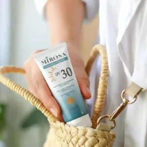 Main tenant un tube de crème solaire MIROSA SPF 30 sortant d'un sac en osier, sur fond de chemise blanche lumineuse.