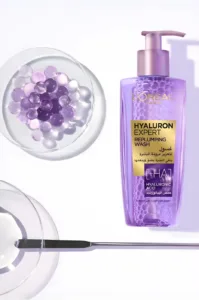 Flacon pompe violet de gel nettoyant Hyaluron Expert à l'acide hyaluronique, posé à côté de perles d'eau et de gel.