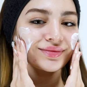 Jeune femme souriante appliquant une texture onctueuse de gel-crème nettoyant sur ses joues pour un soin doux.