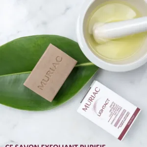 Savon exfoliant Muriac Lightact sur une feuille verte à côté d'un mortier d'huile d'argan et de sa boîte d'emballage.