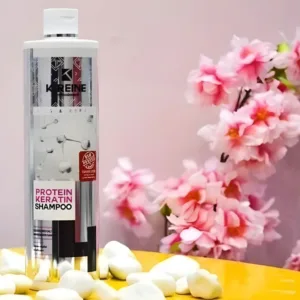 Flacon de shampoing K-Reine Protein Keratin sur table jaune avec galets blancs et fleurs de cerisier roses en fond.