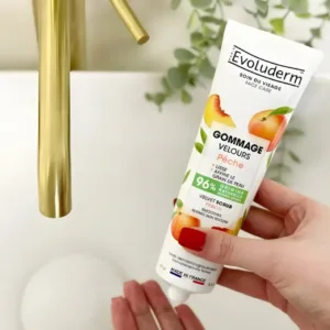Main avec vernis rouge tenant le tube de Gommage Velours Evoluderm au-dessus d'un lavabo blanc avec robinetterie dorée.