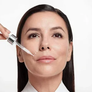 Femme souriante appliquant des gouttes de sérum blanc sur sa joue à l'aide d'une pipette de précision, fond blanc.