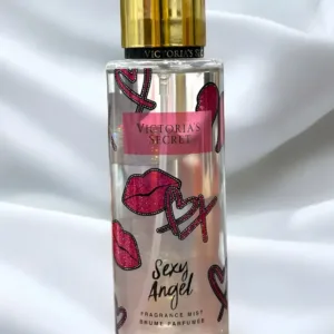 Flacon transparent Brume Victoria's Secret Sexy Angel décoré de lèvres et cœurs roses, bouchon doré sur fond blanc drapé.