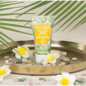 Tube de crème Evoluderm Monoï posé sur un plateau doré avec fleurs de tiaré, galets et feuilles de palmier tropicales.