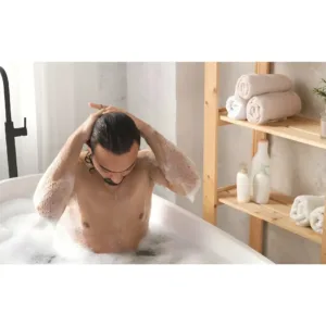 Homme se lavant les cheveux dans un bain moussant, ambiance détente avec étagère en bois, serviettes et soins Dove.