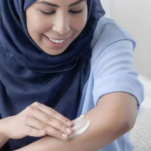 Femme souriante en hijab bleu appliquant la crème hydratante Dove sur son bras. Moment de soin quotidien et bien-être.
