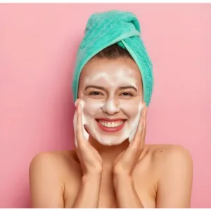 Femme souriante avec serviette turquoise appliquant la texture gel du gommage agrumes sur son visage pour un teint frais.