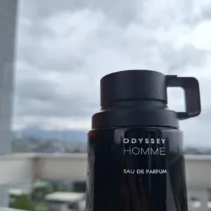 Flacon noir de parfum Odyssey Homme par Armaf posé en extérieur sur un rebord avec vue sur un ciel nuageux et la ville.
